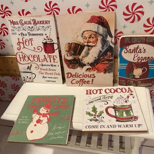 Pode incluir: Cinco placas de madeira de estilo vintage com designs natalinos. As placas apresentam ilustra&ccedil;&otilde;es do Papai Noel, chocolate quente, ponche de ovos e um boneco de neve. As placas s&atilde;o nas cores vermelho, branco e verde e incluem texto como "Chocolate quente", "Ponche de ovos do Papai Noel" e "Chocolate quente".