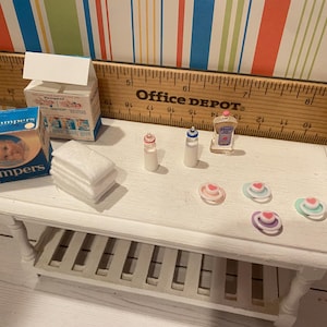 May include: Miniature baby care items on a white table. Includes a Pampers box, baby bottles, a bottle of baby lotion, a stack of white cloths, and several pacifiers. The table is next to a ruler.