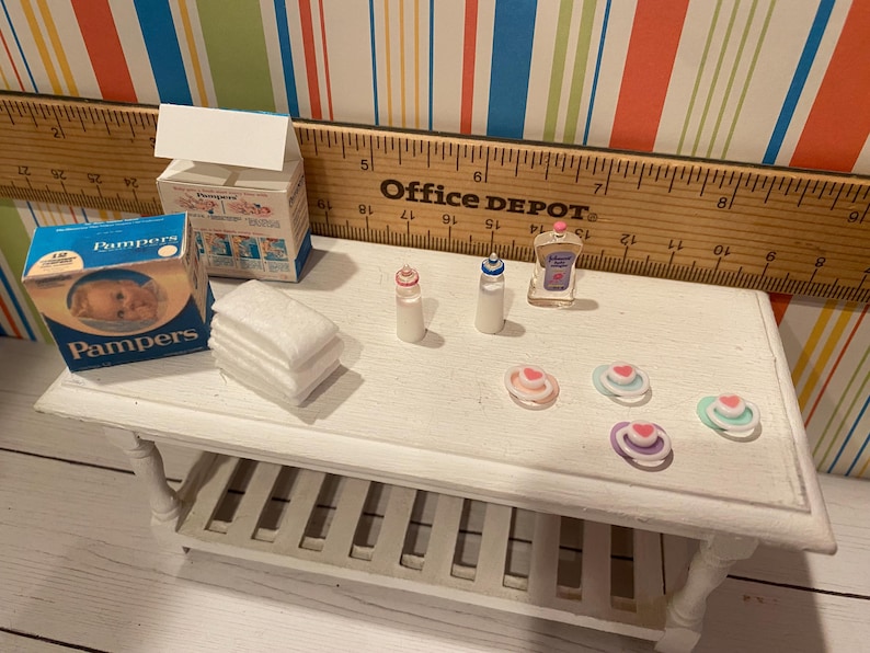 May include: Miniature baby care items on a white table. Includes a Pampers box, baby bottles, a bottle of baby lotion, a stack of white cloths, and several pacifiers. The table is next to a ruler.
