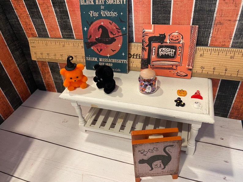 May include: A miniature Halloween scene with an orange teddy bear wearing a witch's hat, a black teddy bear, a glass jar filled with candy, and a sign that says "Boo".