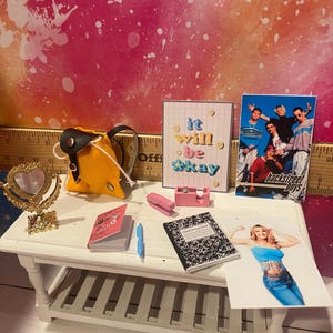 K&ouml;nnte beinhalten: Eine Miniatur-Puppenhausszene mit einem gelben Rucksack, einem rosa Hefter, einem Notizbuch, einem herzf&ouml;rmigen Spiegel und einem gerahmten Druck, auf dem "it will be okay" steht. Es gibt auch ein gerahmtes Foto der Backstreet Boys und ein Foto einer Frau, die ein blaues Oberteil und Jeans tr&auml;gt.
