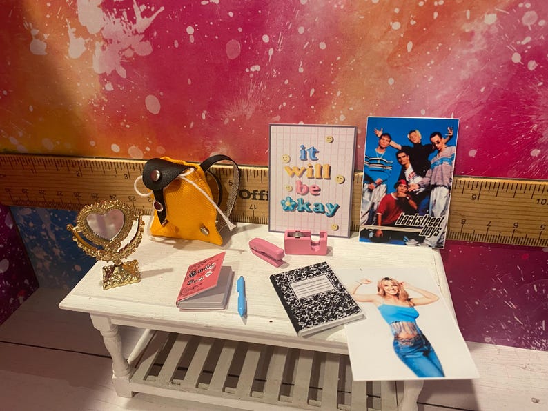 K&ouml;nnte beinhalten: Eine Miniatur-Puppenhausszene mit einem gelben Rucksack, einem rosa Hefter, einem Notizbuch, einem herzf&ouml;rmigen Spiegel und einem gerahmten Druck, auf dem "it will be okay" steht. Es gibt auch ein gerahmtes Foto der Backstreet Boys und ein Foto einer Frau, die ein blaues Oberteil und Jeans tr&auml;gt.