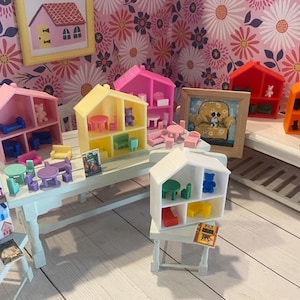 May include: A set of miniature dollhouse furniture, including a yellow, pink, red, orange, and white dollhouse with furniture inside. The dollhouses are on a white table with a book and other miniature furniture.