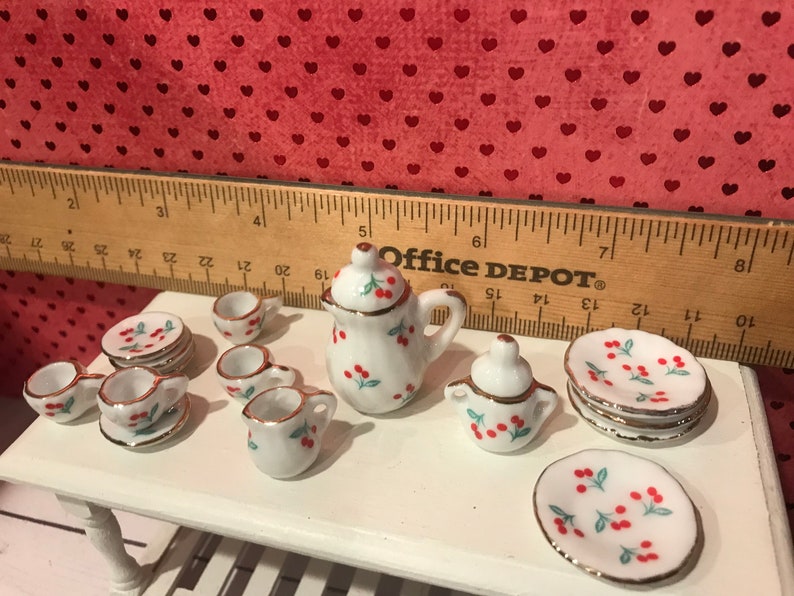 Puede incluir: Un juego de t&eacute; en miniatura con estampado de cerezas que incluye una tetera, una jarra de crema, un azucarero, tazas y platillos. El juego es blanco con cerezas rojas y hojas verdes.