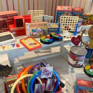 May include: A collection of miniature toys, including board games, a toy oven, and a Lite-Brite set, arranged on a white table. The scene is colorful, with various game boxes and a lion-shaped decoration. A set of colorful hula hoops is in the foreground.