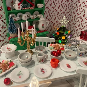 Könnte beinhalten: Miniatur-Weihnachtsszene mit einem weißen Tisch, der mit kleinen Tellern, Schalen und Tassen gedeckt ist. Ein goldener Kerzenleuchter, ein kleiner Weihnachtsbaum und ein Teller mit Keksen stehen auf dem Tisch. Der Hintergrund zeigt ein grünes Regal mit Miniaturgeschirr.