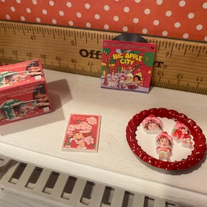Op de afbeelding: Een miniatuursc&egrave;ne met items met het thema Aardbeientaart. Bevat een rode doos met het opschrift "Strawberry Shortcake Garden House", een kleine platenhoes, een boek en een rode schaal met drie Aardbeientaart-beeldjes.