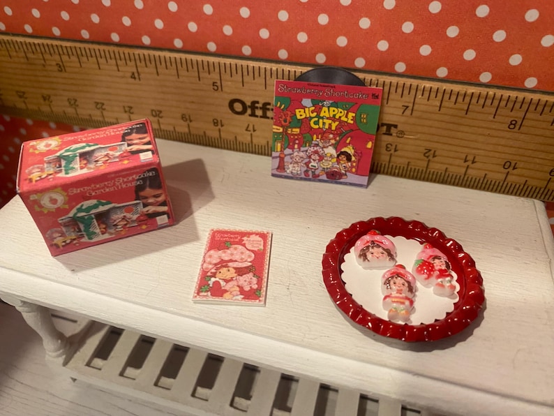 Op de afbeelding: Een miniatuursc&egrave;ne met items met het thema Aardbeientaart. Bevat een rode doos met het opschrift "Strawberry Shortcake Garden House", een kleine platenhoes, een boek en een rode schaal met drie Aardbeientaart-beeldjes.