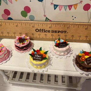 May include: Miniature cakes with colorful frosting and decorations, including a chocolate cake, a yellow cake, and a white cake. Each cake has a "Happy Birthday" sign. The cakes are on a white table.