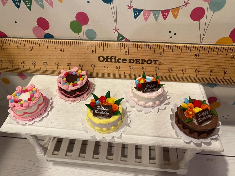 May include: Miniature cakes with colorful frosting and decorations, including a chocolate cake, a yellow cake, and a white cake. Each cake has a "Happy Birthday" sign. The cakes are on a white table.