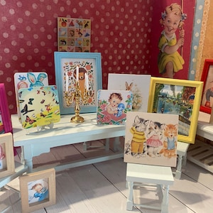 Puede incluir: Una colecci&oacute;n de impresiones art&iacute;sticas enmarcadas de estilo vintage, que incluyen un beb&eacute;, gatitos y una escena de canal. Los marcos son de varios colores, y tambi&eacute;n hay un candelabro dorado. El fondo es una pared rosa con lunares.