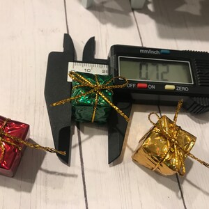 Puede incluir: Tres peque&ntilde;as cajas de regalo envueltas en papel de aluminio verde, dorado y rojo con una cinta dorada. Las cajas se miden con un calibre, que indica 1,8 cm.