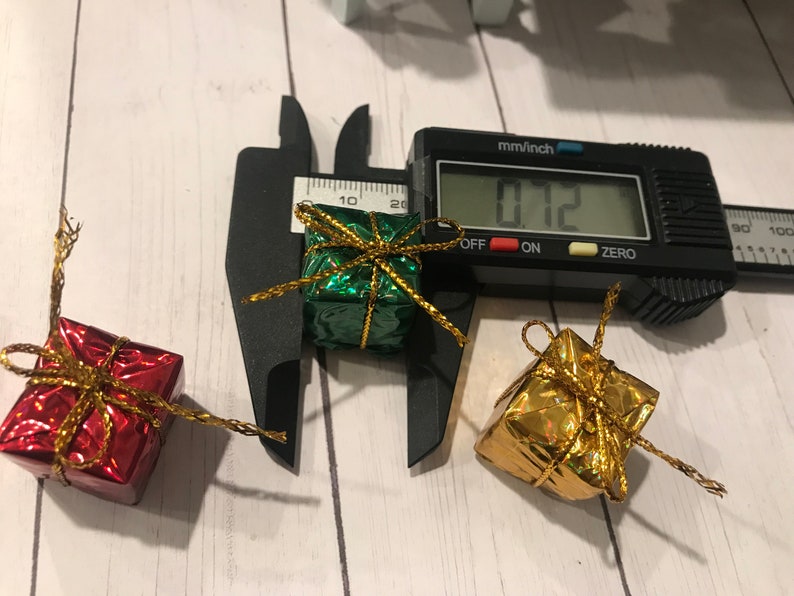 Puede incluir: Tres peque&ntilde;as cajas de regalo envueltas en papel de aluminio verde, dorado y rojo con una cinta dorada. Las cajas se miden con un calibre, que indica 1,8 cm.