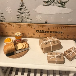 May include: A miniature dollhouse scene with a wooden cutting board, a loaf of bread, a jar of orange jam, a butter knife, and three small brown packages tied with twine. The packages have handwritten addresses and the text "FRONT  C.A." on them. The scene is set on a white table with a wooden ruler in the background.