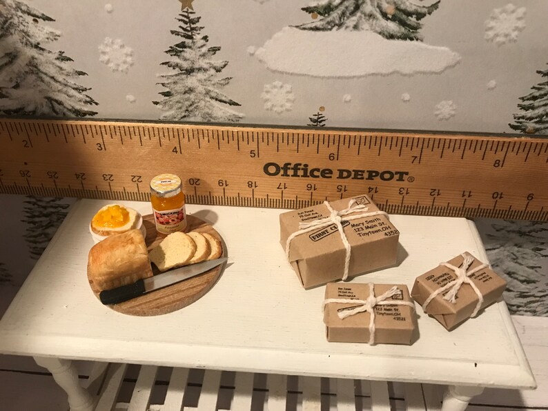 May include: A miniature dollhouse scene with a wooden cutting board, a loaf of bread, a jar of orange jam, a butter knife, and three small brown packages tied with twine. The packages have handwritten addresses and the text "FRONT  C.A." on them. The scene is set on a white table with a wooden ruler in the background.