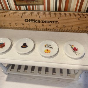 May include: Four small, white, scalloped-edge plates with Thanksgiving-themed designs. One plate features a turkey, another a pilgrim hat, a third says "Happy Thanksgiving" with a pumpkin, and the last has a red maple leaf.