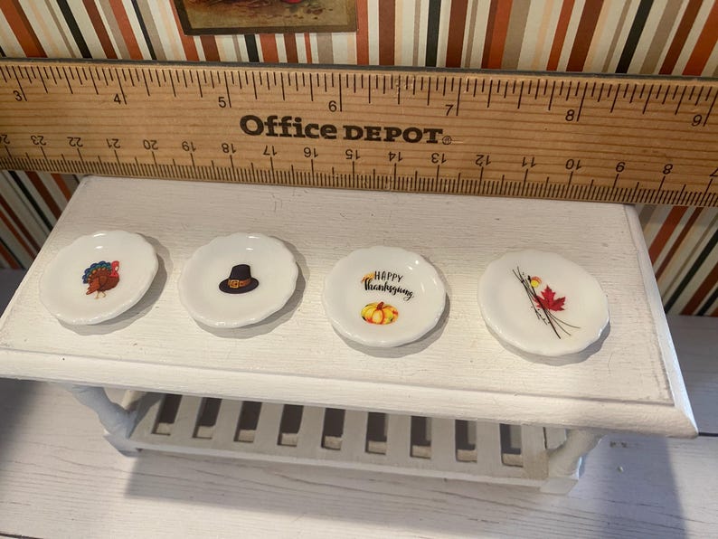May include: Four small, white, scalloped-edge plates with Thanksgiving-themed designs. One plate features a turkey, another a pilgrim hat, a third says "Happy Thanksgiving" with a pumpkin, and the last has a red maple leaf.