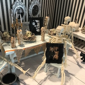 May include: A miniature Halloween scene with skeletons seated at a table. The table is decorated with candles, skulls, and framed vintage photos. A black cat figurine is nearby. The background features a black and white striped wall.