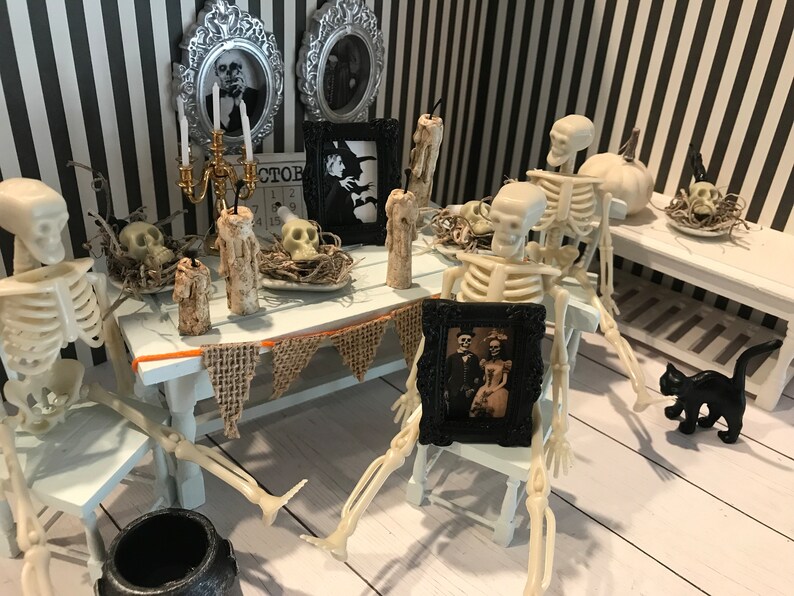 May include: A miniature Halloween scene with skeletons seated at a table. The table is decorated with candles, skulls, and framed vintage photos. A black cat figurine is nearby. The background features a black and white striped wall.