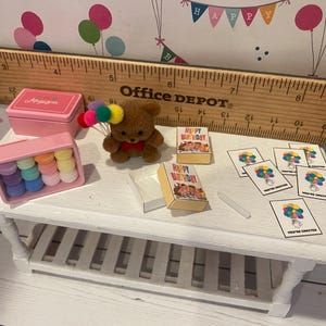 May include: A miniature birthday scene with a brown teddy bear holding colorful balloons. Includes small pink boxes, colorful macarons, miniature books that say "Happy Birthday", and invitation cards. A wooden ruler is in the background.