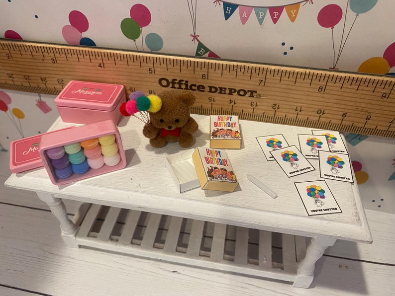 May include: A miniature birthday scene with a brown teddy bear holding colorful balloons. Includes small pink boxes, colorful macarons, miniature books that say "Happy Birthday", and invitation cards. A wooden ruler is in the background.