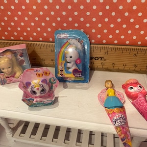 Op de afbeelding: Een verzameling miniatuurspeelgoed, waaronder een pop in een doos, een eenhoornbeeldje en twee Sparkle Girlz kegelvormige speelgoedjes. Het speelgoed is gerangschikt op een witte tafel tegen een achtergrond van een liniaal en een stippenpatroon.