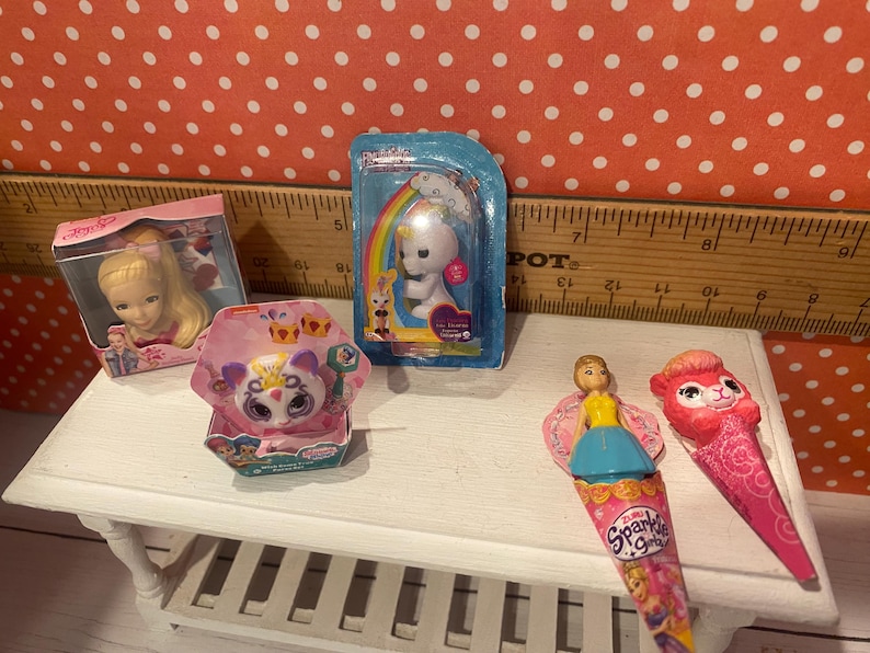 Op de afbeelding: Een verzameling miniatuurspeelgoed, waaronder een pop in een doos, een eenhoornbeeldje en twee Sparkle Girlz kegelvormige speelgoedjes. Het speelgoed is gerangschikt op een witte tafel tegen een achtergrond van een liniaal en een stippenpatroon.