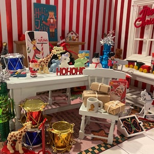 Puede incluir: Una escena navideña en miniatura con una mesa blanca puesta con un tambor rojo y dorado, un cascanueces verde, una figura de jirafa y un letrero que dice "HO HO HO". Hay un caballo de balancín blanco, una silla blanca y una mesa blanca con un fondo rojo y blanco a rayas.
