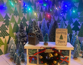 May include: A miniature Christmas scene featuring a white table with a sign that reads "Farm Fresh Christmas Trees Spruce-Pine-Fir". There are several small, snow-covered artificial Christmas trees on the table, along with a wreath and a small metal object.