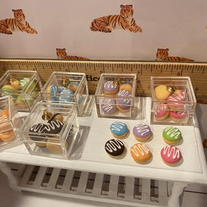 Pode incluir: Macarons e donuts em miniatura em caixas de acr&iacute;lico transparente com bot&otilde;es dourados. Os macarons s&atilde;o em v&aacute;rias cores, incluindo azul, roxo, verde e rosa, com cobertura branca. Os donuts s&atilde;o marrons, laranja e amarelos com cobertura branca.