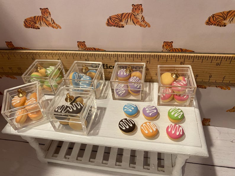 Pode incluir: Macarons e donuts em miniatura em caixas de acr&iacute;lico transparente com bot&otilde;es dourados. Os macarons s&atilde;o em v&aacute;rias cores, incluindo azul, roxo, verde e rosa, com cobertura branca. Os donuts s&atilde;o marrons, laranja e amarelos com cobertura branca.