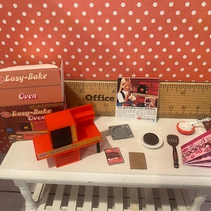 Op de afbeelding: Een miniatuur Easy-Bake Oven set met een oranje oven, een doos en accessoires. De set bevat een kookboek, spatel en andere kleine items, allemaal gerangschikt op een witte tafel. De achtergrond is een rood-wit stippenpatroon.