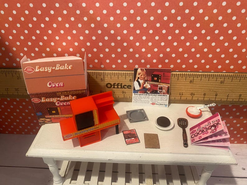 Op de afbeelding: Een miniatuur Easy-Bake Oven set met een oranje oven, een doos en accessoires. De set bevat een kookboek, spatel en andere kleine items, allemaal gerangschikt op een witte tafel. De achtergrond is een rood-wit stippenpatroon.