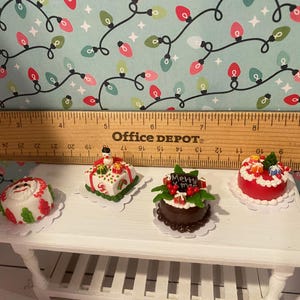 May include: Four miniature Christmas cakes on a white table. The cakes are decorated with red, green, and white frosting and have various Christmas-themed decorations, including a snowman, a Santa hat, and a "Merry Christmas" sign.
