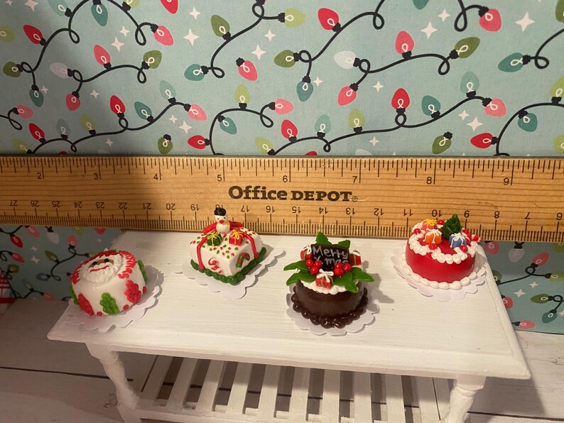 May include: Four miniature Christmas cakes on a white table. The cakes are decorated with red, green, and white frosting and have various Christmas-themed decorations, including a snowman, a Santa hat, and a "Merry Christmas" sign.