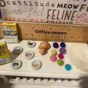 May include: A miniature cat food box with the text "Meow Mix" on it, a miniature cat figurine, five miniature cat food bowls, and a white fluffy cat bed. There are also colorful felt balls scattered around the items.