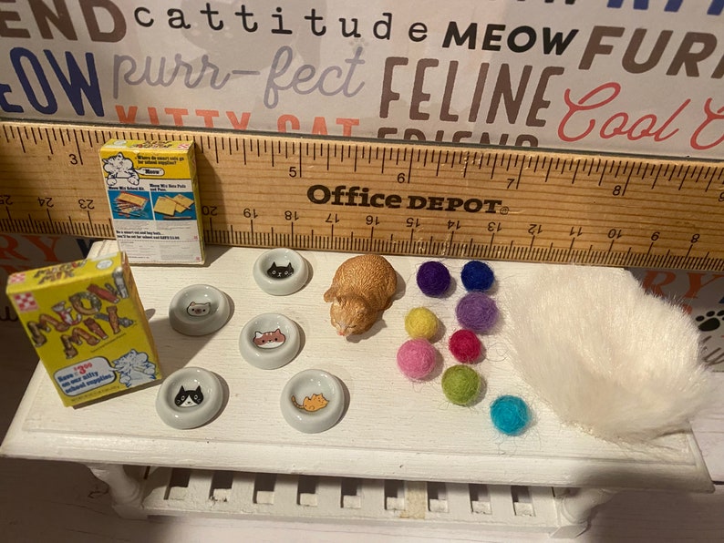 May include: A miniature cat food box with the text "Meow Mix" on it, a miniature cat figurine, five miniature cat food bowls, and a white fluffy cat bed. There are also colorful felt balls scattered around the items.