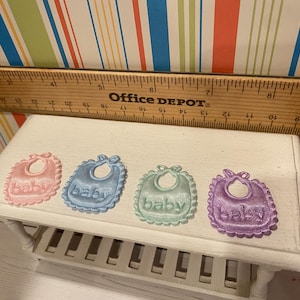 May include: Four pastel-colored baby bibs with the word "baby" embroidered on them. The bibs are pink, blue, green, and purple, each with a scalloped edge. A wooden ruler is placed above the bibs.
