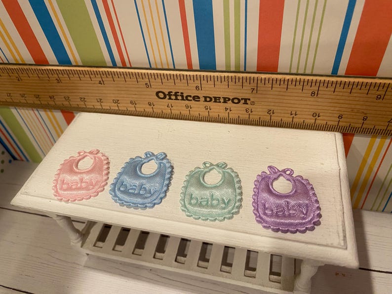 May include: Four pastel-colored baby bibs with the word "baby" embroidered on them. The bibs are pink, blue, green, and purple, each with a scalloped edge. A wooden ruler is placed above the bibs.