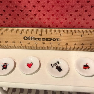 Puede incluir: Cuatro peque&ntilde;os platos de cer&aacute;mica blanca con dise&ntilde;os en negro y rojo. Los platos presentan un oso con alas, un coraz&oacute;n, un oso con la palabra "Love" y un oso conduciendo un coche rojo.