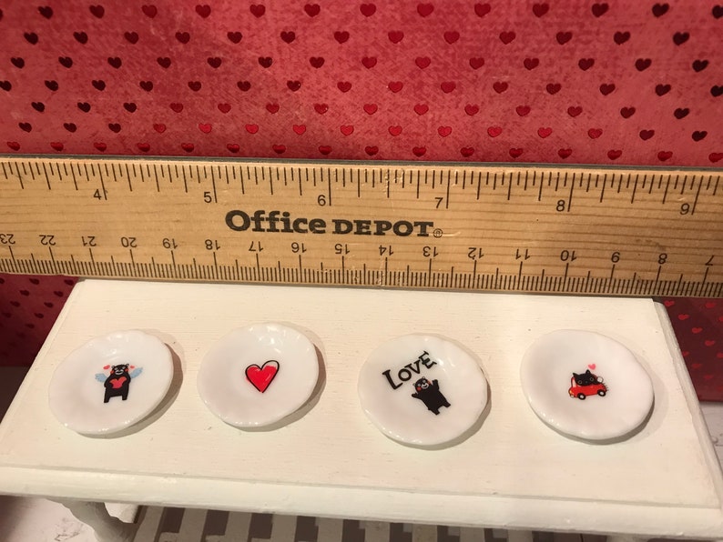 Puede incluir: Cuatro peque&ntilde;os platos de cer&aacute;mica blanca con dise&ntilde;os en negro y rojo. Los platos presentan un oso con alas, un coraz&oacute;n, un oso con la palabra "Love" y un oso conduciendo un coche rojo.
