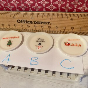 May include: Three small, white, decorative plates with Christmas-themed designs. One plate features a Christmas tree, another a snowman, and the third Santa Claus in a sleigh with reindeer. A wooden ruler is in the background.