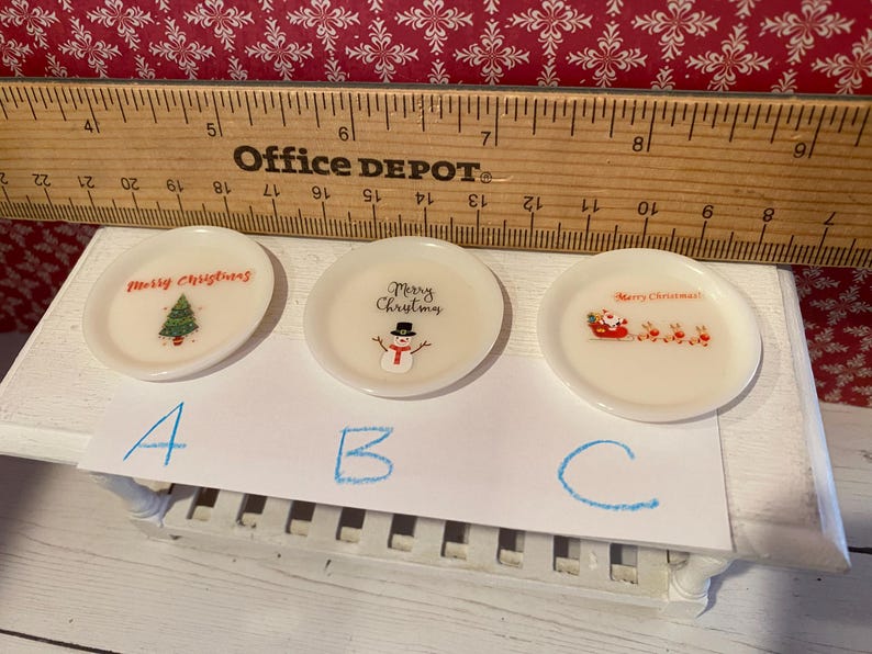 May include: Three small, white, decorative plates with Christmas-themed designs. One plate features a Christmas tree, another a snowman, and the third Santa Claus in a sleigh with reindeer. A wooden ruler is in the background.