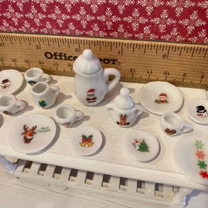 May include: A miniature Christmas-themed tea set. The white porcelain set includes a teapot, creamer, sugar bowl, cups, and saucers. Each piece is decorated with festive images of Santa, snowmen, reindeer, and Christmas trees.