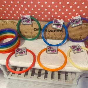 Op de afbeelding: Een verzameling kleurrijke hoelahoeps in verschillende tinten, waaronder rood, oranje, geel, groen, blauw en paars. Elke hoepel heeft een klein label met de tekst "hula hoop". De hoepels zijn gerangschikt op een witte tafel met een houten liniaal op de achtergrond.