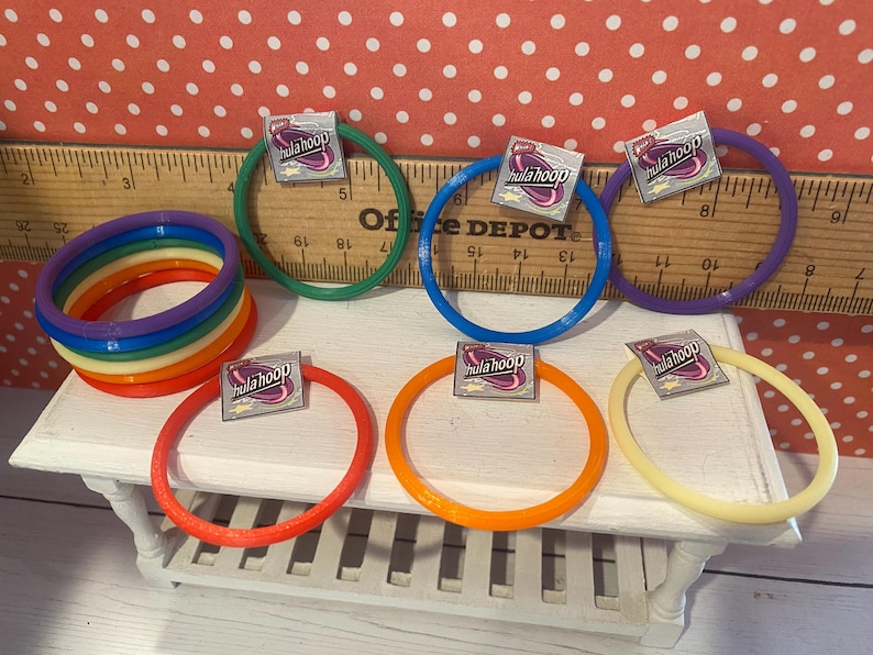 Op de afbeelding: Een verzameling kleurrijke hoelahoeps in verschillende tinten, waaronder rood, oranje, geel, groen, blauw en paars. Elke hoepel heeft een klein label met de tekst "hula hoop". De hoepels zijn gerangschikt op een witte tafel met een houten liniaal op de achtergrond.