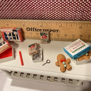 May include: Miniature Christmas-themed items, including boxes of Noma Lites Candolier, ornaments, and gingerbread baking mix. A gingerbread man with a red bow tie and blue buttons is also present. A red candle and a small metal screw are also visible.