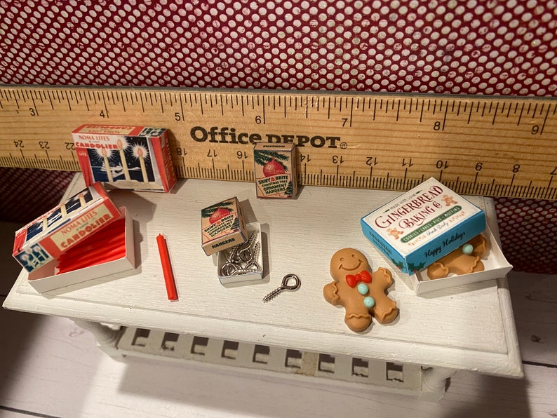 May include: Miniature Christmas-themed items, including boxes of Noma Lites Candolier, ornaments, and gingerbread baking mix. A gingerbread man with a red bow tie and blue buttons is also present. A red candle and a small metal screw are also visible.