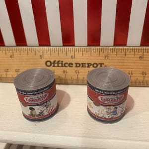 May include: Two miniature toy cans with gray lids and red and white labels. The labels read "The Classic Tinkertoy Ultra Construction Set" with illustrations of children playing. A wooden ruler is behind the cans.