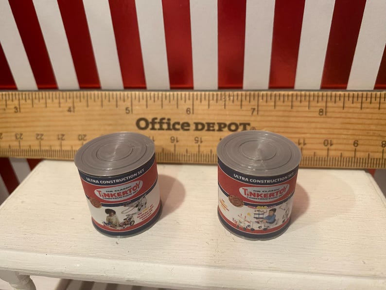 May include: Two miniature toy cans with gray lids and red and white labels. The labels read "The Classic Tinkertoy Ultra Construction Set" with illustrations of children playing. A wooden ruler is behind the cans.